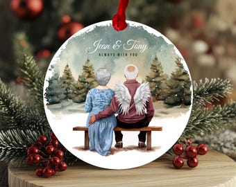 May include: A round, white ornament with a watercolor painting of a couple sitting on a bench in a snowy forest. The text "Jean & Tony Always With You" is written on the ornament.