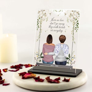 May include: A white acrylic memorial plaque with a floral design and the text "Those we love don't go away they walk beside us everyday". Two figures are depicted, one with angel wings, sitting on a bench. The plaque has a black base and the names "Laura & Mom" are written below the figures.
