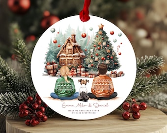 May include: A round Christmas ornament with a watercolor illustration of a couple and their dog standing in front of a decorated Christmas tree and a gingerbread house. The text "Emma, Milo & Daniel" and "When we have each other we have everything" is written below the illustration.