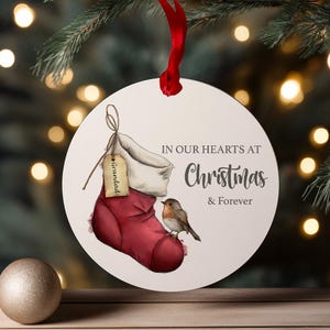 May include: A circular Christmas tree decoration with a red ribbon. It depicts a red stocking with a "Grandad" tag, a robin, and the words "IN OUR HEARTS AT Christmas & Forever".