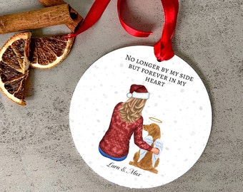 May include: A white round ornament with a red ribbon. The ornament features a woman in a red sweater and a Santa hat, holding a dog with angel wings. The text on the ornament reads "No longer by my side but forever in my heart" and "Lara & Max".