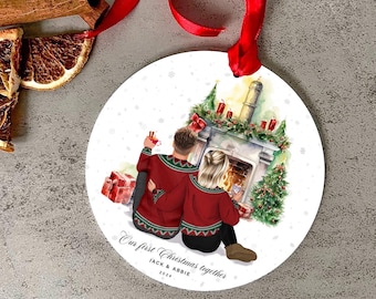 May include: A white round ornament with a watercolor illustration of a couple in red sweaters sitting by a fireplace. The text "Our first Christmas together" is written below the couple, along with the names "Jack & Abbie" and the year "2024".
