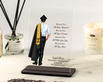 May include: A clear acrylic plaque with a black base featuring a graduate in a black cap and gown with a gold sash. The text reads "Behind You All Your Memories Before You All Your Dreams Around You All Who Love You Within You All You Need Sajid Khan Manchester Metropolitan University BA (Hons) Business".