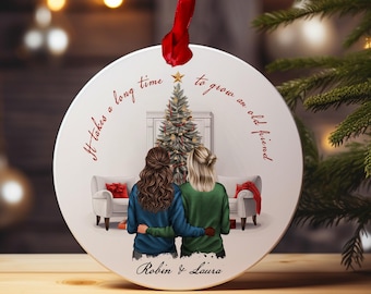 Pode incluir: Um enfeite de Natal redondo branco com uma árvore de Natal e duas mulheres em pé na frente dela. O enfeite tem o texto "Leva muito tempo para se tornar um velho amigo" e "Robin & Laura".