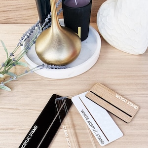 May include: Three different display stands for products. A black acrylic stand, a clear acrylic stand, and a wood stand. Each stand has the name of the stand printed on it in white text.