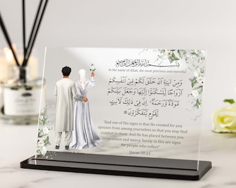 May include: A white glass plaque with a black frame features an illustration of a couple in traditional Islamic clothing. The plaque includes an Arabic inscription and a translation in English, "And one of His signs is that He created for you spouses from among yourselves so that you may find comfort in them. And He has placed between you compassion and mercy. Surely in this are signs for people who reflect." The plaque also includes the Quran verse 30:21 and the names "Omar & Aliya".