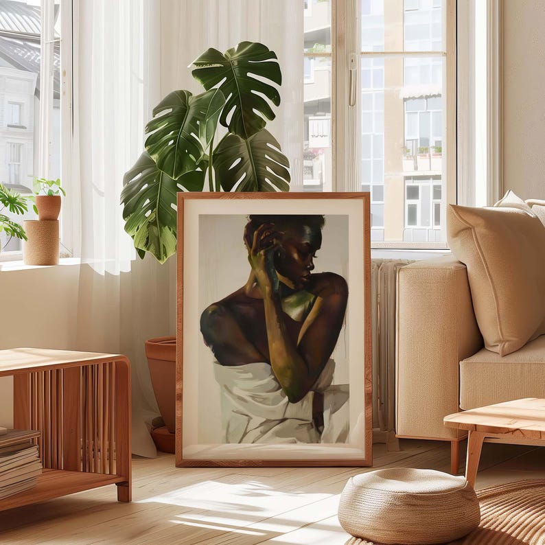 May include: A framed art print featuring a portrait of a person with dark skin, draped in white fabric. The artwork is set in a wooden frame and displayed in a well-lit living room with a large plant and wooden furniture.