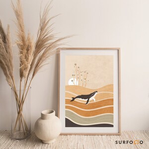 May include: A framed art print featuring a minimalist illustration of a whale swimming in a sea of layered, earthy tones. The artwork includes a sun and stylized plants against a beige background. The print is in a light wood frame.