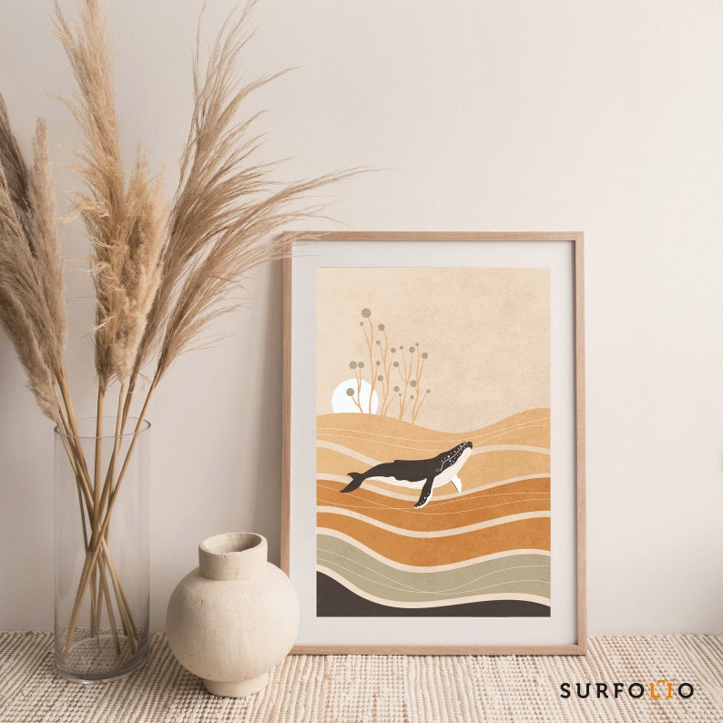 May include: A framed art print featuring a minimalist illustration of a whale swimming in a sea of layered, earthy tones. The artwork includes a sun and stylized plants against a beige background. The print is in a light wood frame.