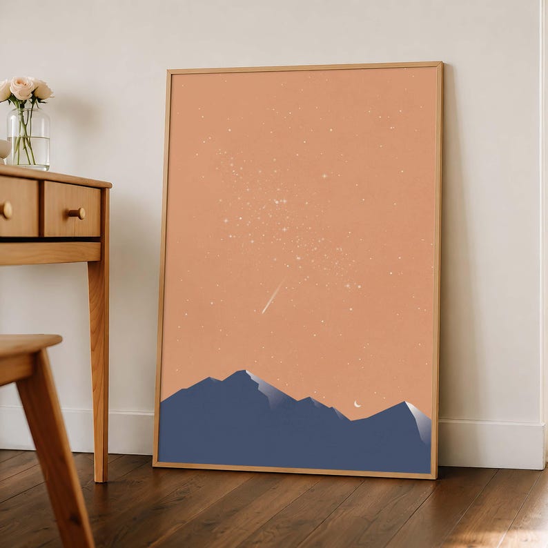 May include: A rectangular framed wall art print featuring a dark blue mountain range silhouette against a solid peach-colored sky. The sky is filled with scattered white stars and a single shooting star, with a small white crescent moon visible.