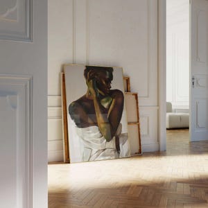 May include: An oil painting of a person with dark skin, draped in white fabric, leaning against a wall. The artwork is in a gold frame and is leaning against other canvases. The room has white walls and a wooden floor.