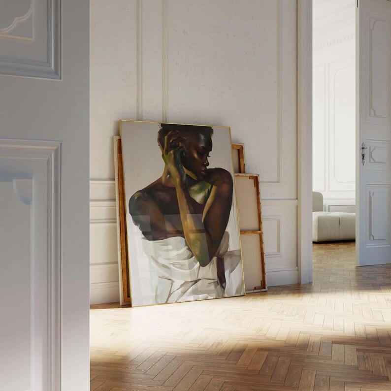 May include: An oil painting of a person with dark skin, draped in white fabric, leaning against a wall. The artwork is in a gold frame and is leaning against other canvases. The room has white walls and a wooden floor.