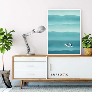 May include: A white framed print of two people on surfboards in a blue ocean with white waves. The print is hanging on a white wall above a white dresser with three drawers. The dresser has a wooden top and legs. A lamp is on the left side of the dresser. The text "SURFOLO" is on the front of the dresser.