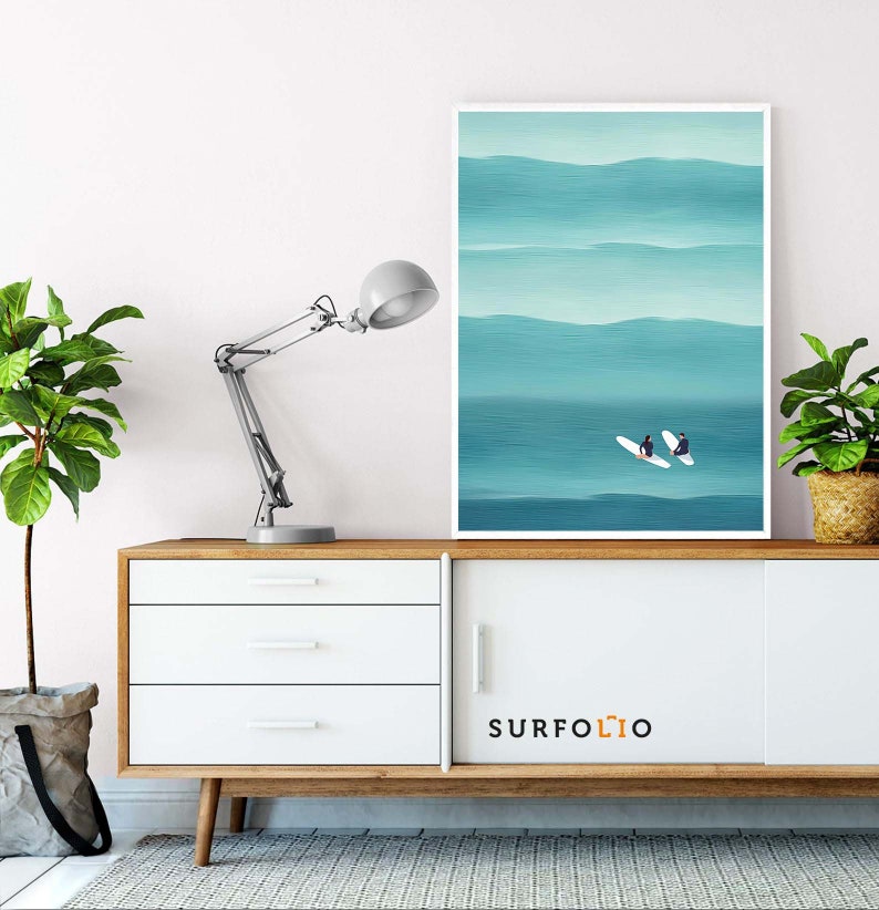 May include: A white framed print of two people on surfboards in a blue ocean with white waves. The print is hanging on a white wall above a white dresser with three drawers. The dresser has a wooden top and legs. A lamp is on the left side of the dresser. The text "SURFOLO" is on the front of the dresser.
