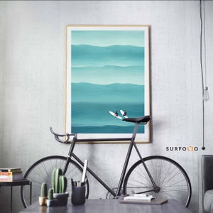 May include: A framed abstract painting of a turquoise ocean with two surfers riding waves. The painting is hanging on a white wall above a black bicycle.