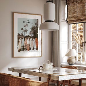 May include: A framed print with the text "THE COASTAL" above a row of surfboards on a beach. The image features palm trees and a vintage aesthetic. The frame is a light brown color.