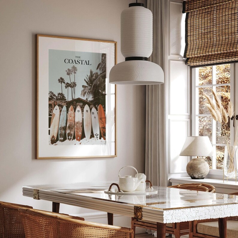 May include: A framed print with the text "THE COASTAL" above a row of surfboards on a beach. The image features palm trees and a vintage aesthetic. The frame is a light brown color.