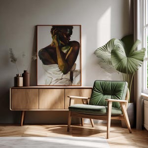 May include: A framed artwork depicting a person draped in white fabric, displayed above a wooden cabinet. A green armchair with a wooden frame sits nearby, complemented by a large potted plant and a window with natural light.
