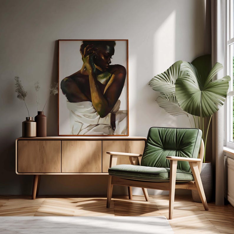 May include: A framed artwork depicting a person draped in white fabric, displayed above a wooden cabinet. A green armchair with a wooden frame sits nearby, complemented by a large potted plant and a window with natural light.