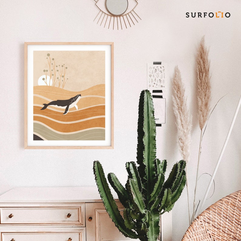 May include: A framed art print featuring a minimalist illustration of a whale swimming in a sea of layered, earthy tones. The artwork has a light wood frame and a neutral color palette, with a sun and plant details in the background. The print is labeled with the word "SURFOLIO".