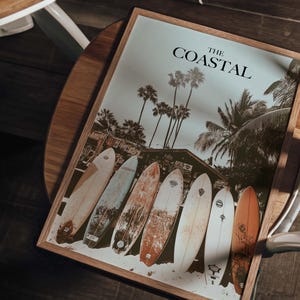 May include: A framed print with the text "THE COASTAL" features a beach scene with palm trees and surfboards. The image shows six surfboards of various colors and designs leaning against the sand.