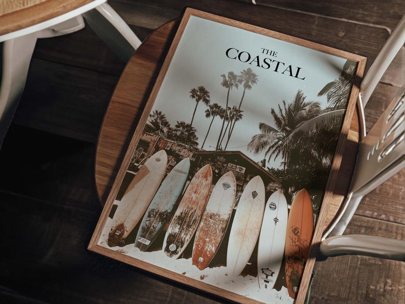 May include: A framed print with the text "THE COASTAL" features a beach scene with palm trees and surfboards. The image shows six surfboards of various colors and designs leaning against the sand.