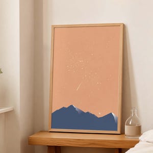May include: A vertical rectangular framed wall art print featuring a dark blue mountain range silhouette against a solid peach-colored sky. The sky is filled with scattered white stars and a single shooting star, with a small white crescent moon.