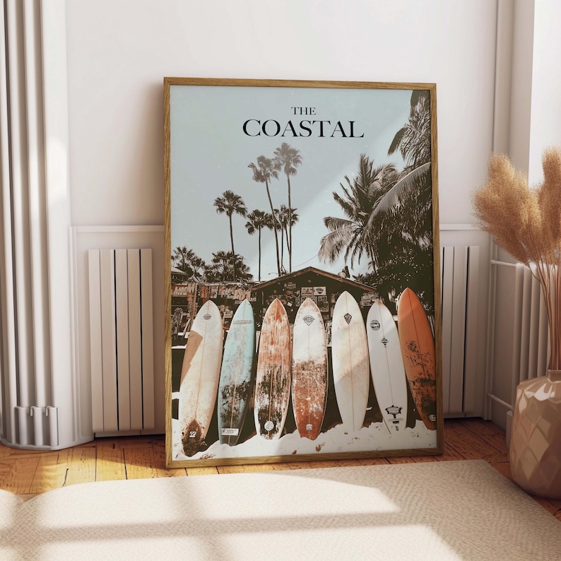 May include: Framed print featuring the text "THE COASTAL" above a beach scene with palm trees and surfboards. Seven surfboards of various colors and designs are lined up in front of a beach shack. The print is in a wooden frame.