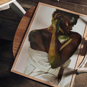 May include: A framed art print featuring a portrait of a person with dark skin, draped in white fabric. The artwork has a warm color palette with shades of brown, green, and white. The frame is made of light-colored wood, and the print is resting on a wooden surface.