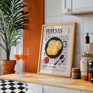 May include: A framed print with the word "PUPUSAS" at the top, featuring an illustration of pupusas, a Salvadoran dish. The print is in a gold frame and is displayed on a wooden surface. Two wine glasses with orange liquid are in front of the print.