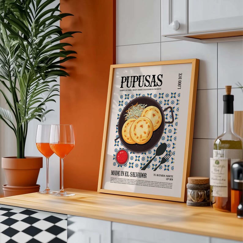 May include: A framed print with the word "PUPUSAS" at the top, featuring an illustration of pupusas, a Salvadoran dish. The print is in a gold frame and is displayed on a wooden surface. Two wine glasses with orange liquid are in front of the print.