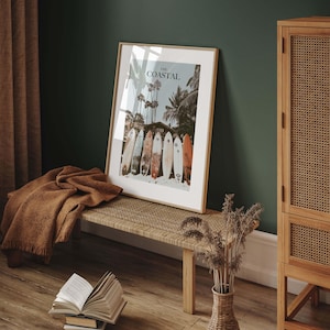 May include: Framed print with the text "The Coastal" featuring a beach scene with palm trees and surfboards. The print is displayed on a woven bench with a brown blanket and a vase of dried flowers. A wooden cabinet is on the right.