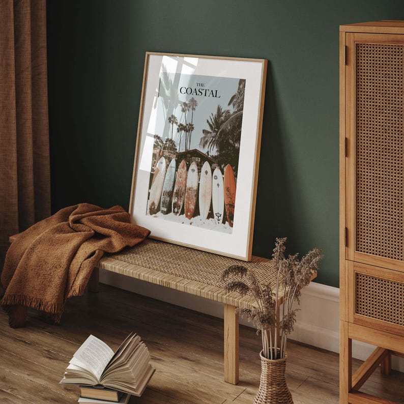 May include: Framed print with the text "The Coastal" featuring a beach scene with palm trees and surfboards. The print is displayed on a woven bench with a brown blanket and a vase of dried flowers. A wooden cabinet is on the right.