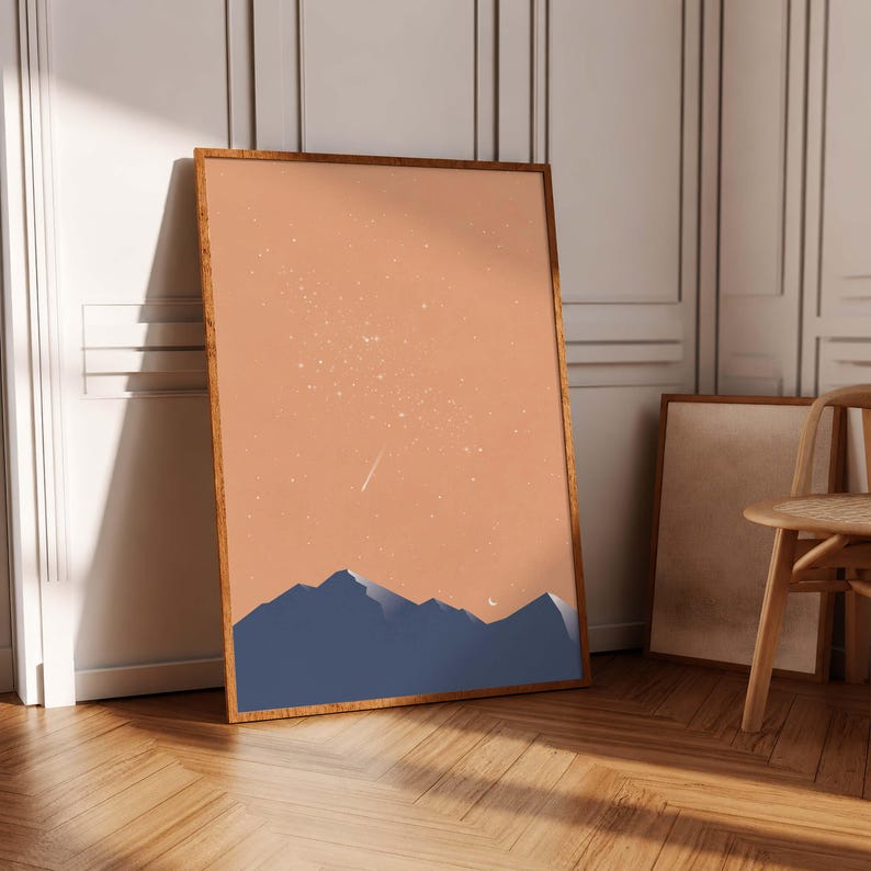 May include: A rectangular framed wall art print featuring a dark blue mountain range silhouette against a solid peach-colored sky filled with white stars and a small crescent moon. The artwork is encased in a simple, thin wooden frame.