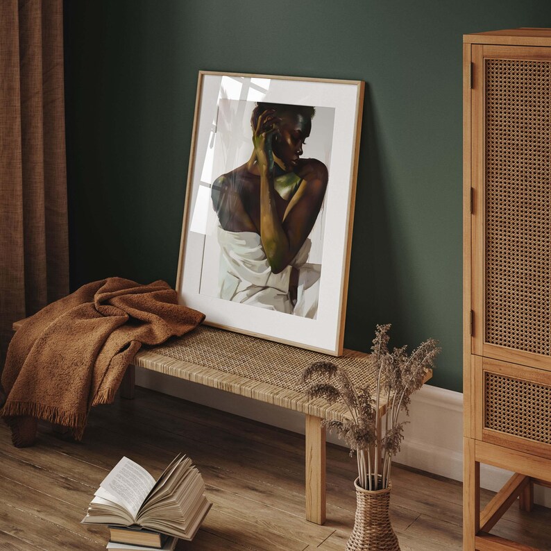 May include: A framed art print featuring a portrait of a person wrapped in a white cloth, with green accents. The artwork is displayed on a woven bench, alongside a brown blanket, books, and a vase with dried plants. The room has a dark green wall.