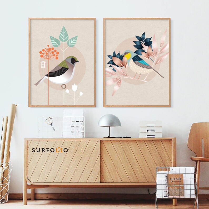 May include: Two framed prints with a minimalist design featuring stylized birds and floral elements. The prints have a light beige background with pink, blue, and green accents. The prints are displayed on a wooden shelf with a lamp and other decorative items.