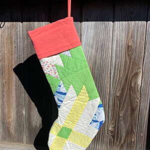 May include: A patchwork Christmas stocking with a green, yellow, and blue design. The stocking has a coral red cuff and is hanging from a hook.