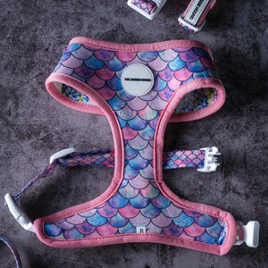 May include: A pink and mermaid scale patterned dog harness with matching collar and leash. The harness has a pink trim and white buckles. The text "THE AUSSIE HOUND" is printed on the collar and harness.