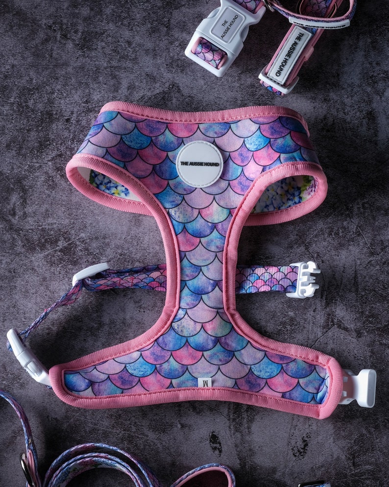 May include: A pink and mermaid scale patterned dog harness with matching collar and leash. The harness has a pink trim and white buckles. The text "THE AUSSIE HOUND" is printed on the collar and harness.