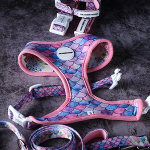 May include: A dog harness and leash set with a mermaid scale pattern in shades of pink, purple, and blue. The harness has a pink trim and white plastic buckles. The leash and harness have the text "THE AUSSIE HOUND".