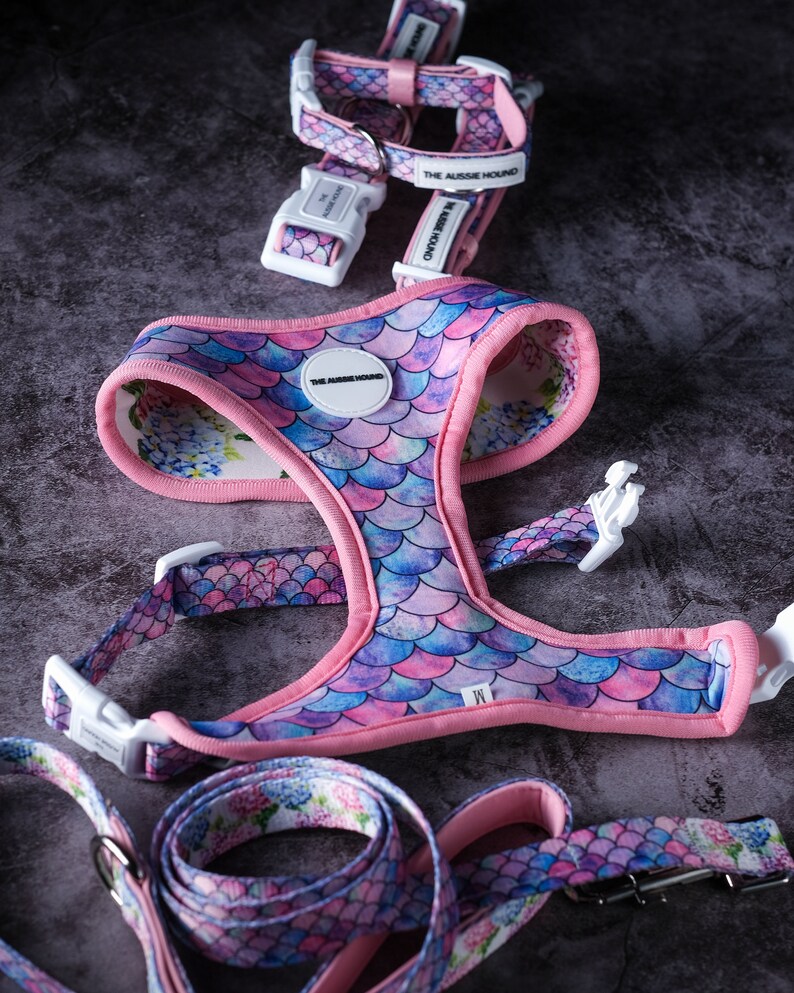 May include: A dog harness and leash set with a mermaid scale pattern in shades of pink, purple, and blue. The harness has a pink trim and white plastic buckles. The leash and harness have the text "THE AUSSIE HOUND".