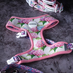 May include: A dog harness, collar, and leash set featuring a floral hydrangea pattern in pink, purple, and blue on a white background. The harness and collar have pink trim and white plastic buckles. The text "THE AUSSIE HOUND" is visible on the collar.