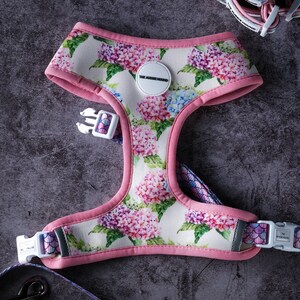 May include: A dog harness with a floral hydrangea pattern in pink, purple, and blue on a white background. The harness has pink trim and a white buckle. The brand name "THE AUSSIE HOUND" is printed on a white circular patch.