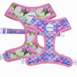 May include: Two dog harnesses. One features a floral pattern with pink, purple, and blue flowers on a cream background. The other has a mermaid scale design in shades of pink, purple, and blue. Both have pink trim and white buckles.
