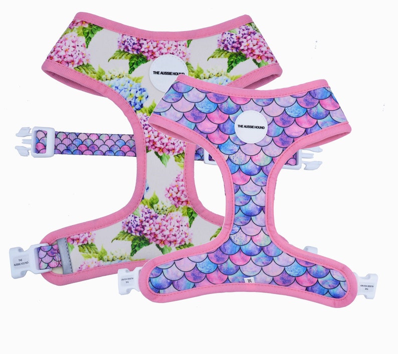 May include: Two dog harnesses. One features a floral pattern with pink, purple, and blue flowers on a cream background. The other has a mermaid scale design in shades of pink, purple, and blue. Both have pink trim and white buckles.