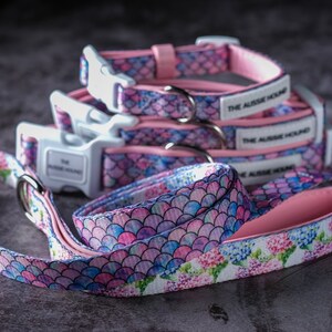Op de afbeelding: Een set hondenhalsbanden en een bijpassende riem met een zeemeerminstaartpatroon in roze, blauw en paars. De halsbanden hebben witte plastic gespen en de tekst "THE AUSSIE HOUND". De riem heeft een bloemenpatroon met roze voering.