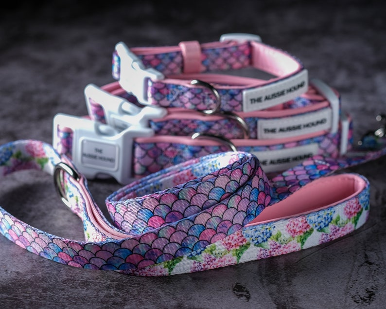 Op de afbeelding: Een set hondenhalsbanden en een bijpassende riem met een zeemeerminstaartpatroon in roze, blauw en paars. De halsbanden hebben witte plastic gespen en de tekst "THE AUSSIE HOUND". De riem heeft een bloemenpatroon met roze voering.