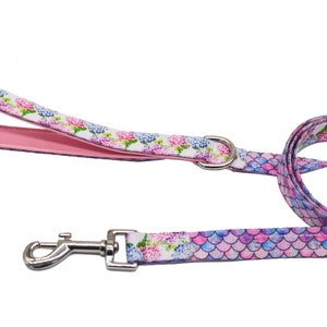 Pode incluir: Trela para cães com um padrão floral e de escamas de sereia. A trela apresenta um fundo branco com flores rosa, azuis e roxas. A pega é rosa com um padrão floral. Uma fivela de metal prateado está presa.