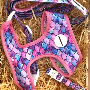 May include: A dog harness, leash, and collar set in a wooden box. The harness and collar feature a mermaid scale pattern in shades of pink, blue, and purple, with pink trim. The leash has a floral pattern. The set includes the text "THE AUSSIE HOUND".