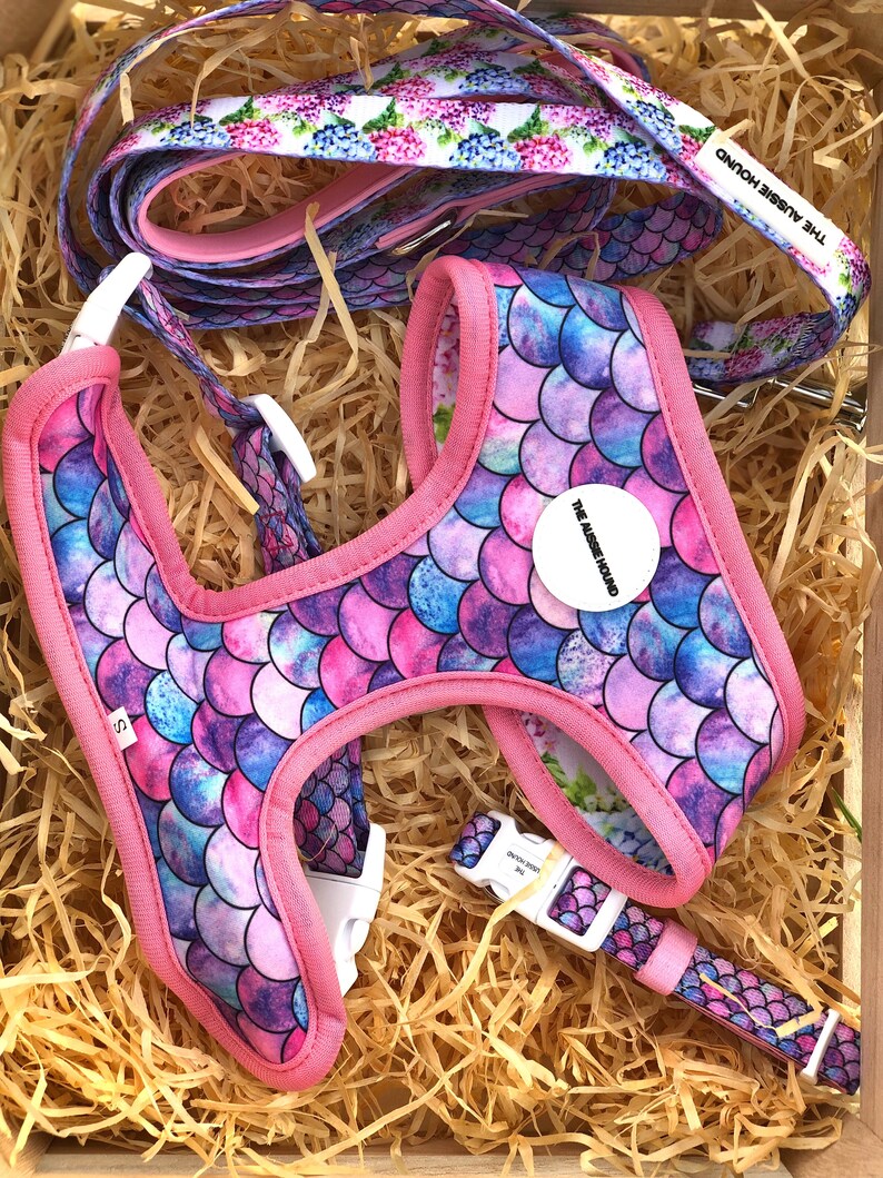 May include: A dog harness, leash, and collar set in a wooden box. The harness and collar feature a mermaid scale pattern in shades of pink, blue, and purple, with pink trim. The leash has a floral pattern. The set includes the text "THE AUSSIE HOUND".